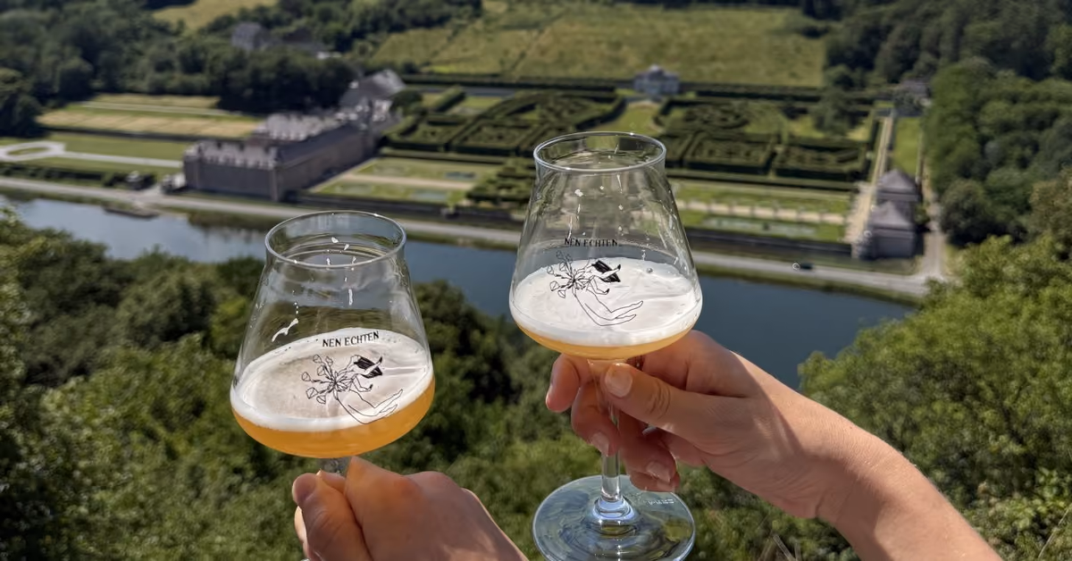 Two glasses of beer raised above the formal gardens of Château de Freÿr and the river Meuse